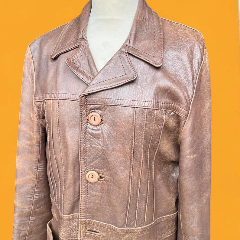 May include: A brown leather jacket featuring a pointed collar, long sleeves, and a front button closure with two visible round buttons. The garment displays vertical stitched paneling details across the chest and a textured finish throughout.