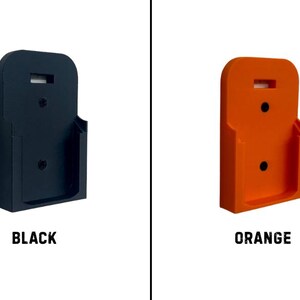 May include: Two wall-mounted holders, one black and one orange, are displayed against a white background. Each holder has a rectangular shape with a curved top and a small shelf. The word "BLACK" is below the black holder, and "ORANGE" is below the orange holder.