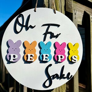 Może przedstawiać: Okrągły, biały drewniany znak z napisem "Oh For Peeps Sake" napisanym czarnym pismem. Pięć kolorowych drewnianych figurek piskląt ułożonych jest w rzędzie pod napisem, każda z białą literą tworzącą słowo "PEEPS".