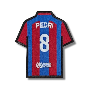 Può includere: Maglia da calcio a strisce rosse e blu con il nome "PEDRI" e il numero "8" sul retro. La maglia ha un logo UNHCR in basso.