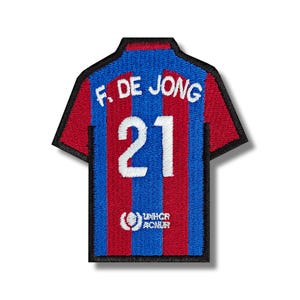 Può includere: Toppa ricamata a forma di maglia da calcio. La maglia è a righe blu e rosse, con il nome "F. DE JONG" e il numero "21" in bianco. La toppa presenta anche il logo UNHCR/ACNUR.