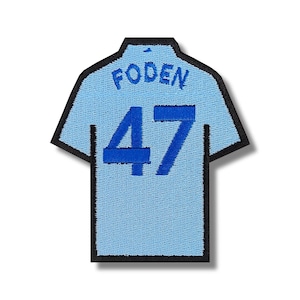 May include: A light blue embroidered patch shaped like a soccer jersey with the name "Foden" and the number "47" in blue.