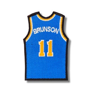 Pode incluir: Um patch de camisa azul com o nome "BRUNSON" e o número "11" em amarelo. A camisa tem uma borda preta com detalhes em amarelo e branco. O patch foi projetado para se assemelhar a uma camisa de basquete.