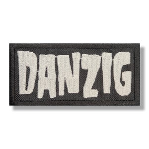 Może przedstawiać: Haftowana naszywka z napisem "DANZIG" w pogrubionych, białych literach. Tekst jest wyszyty na czarnym prostokątnym tle z czarną obwódką. Naszywka ma czysty, profesjonalny wygląd.