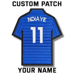 May include: A blue embroidered patch in the shape of a soccer jersey. The jersey has the name "NDIAYE" and the number "11" in white. The text "CUSTOM PATCH" and "YOUR NAME" are above and below the jersey.