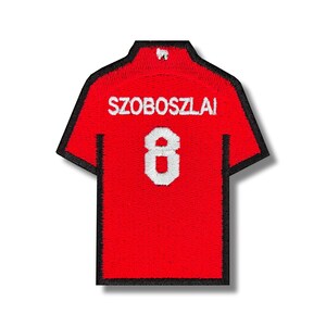 Könnte beinhalten: Roter Fußballtrikot-Patch mit schwarzem Besatz, mit dem Namen "SZOBOSZLAI" und der Nummer "8" in Weiß.