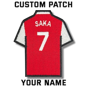 May include: A custom patch in the shape of a red and white soccer jersey. The jersey features the name "SAKA" and the number "7" in white. The text "CUSTOM PATCH" and "YOUR NAME" are printed above and below the jersey.
