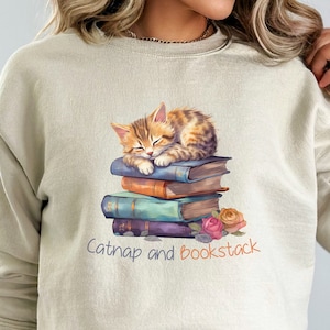 May include: A beige sweatshirt with a graphic of a cat napping on a stack of books. The text "Catnap and Bookstack" is printed below the image.