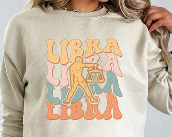 May include: A light beige sweatshirt with a graphic design of the word "LIBRA" repeated in different colors and a stylized image of a person holding a scale.