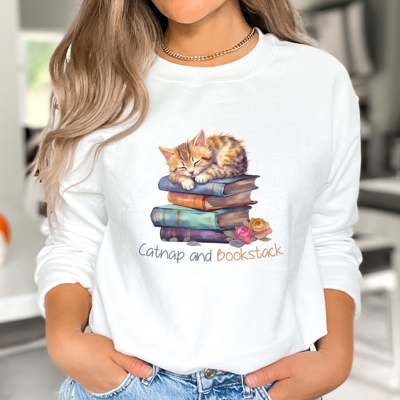 May include: White sweatshirt with a graphic of a cat napping on a stack of books. The text "Catnap and Bookstack" is printed below the image.