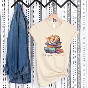 May include: A beige t-shirt with a graphic of an orange tabby cat napping on a stack of colorful books. The text "Catnap and Bookstack" is printed below the cat.