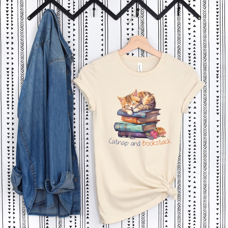 May include: A beige t-shirt with a graphic of an orange tabby cat napping on a stack of colorful books. The text "Catnap and Bookstack" is printed below the cat.