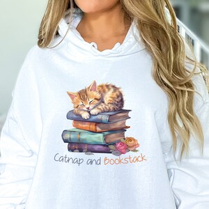 May include: White hoodie with a cute orange and brown tabby cat sleeping on a stack of colorful books. The text "Catnap and Bookstack" is printed below the cat.