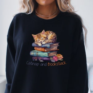 May include: A black sweatshirt with a cute illustration of a tabby cat napping on a stack of colorful books. The text "Catnap and Bookstack" is printed below the image.