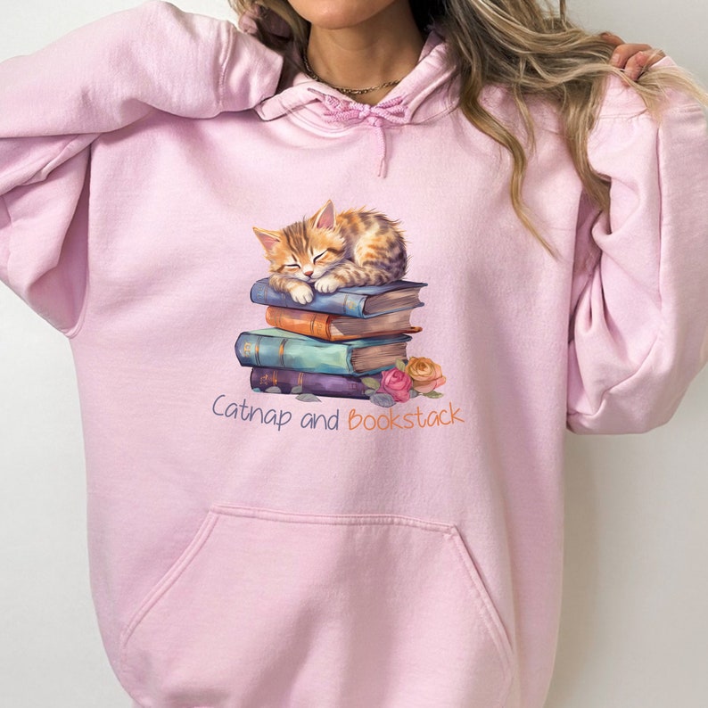 May include: Pink hooded sweatshirt with a graphic of a cat napping on a stack of books. The text "Catnap and Bookstack" is printed below the image.
