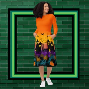 May include: An orange long-sleeved dress with a Halloween-themed skirt. The skirt features a spooky house design with pumpkins, a fence, and a bat. The dress has pockets and is knee-length.