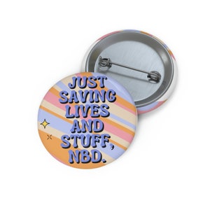 Może przedstawiać: Okrągła przypinka z agrafką. Przypinka ma pastelową kolorystykę z ukośnymi paskami w kolorze pomarańczowym, różowym i niebieskim. Na przodzie nadrukowano tekst "JUST SAVING LIVES AND STUFF, NBD." z małą grafiką gwiazdy.