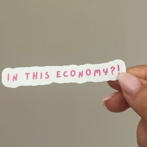 May include: A white sticker with the phrase "IN THIS ECONOMY?!" in pink handwritten font. The sticker has a torn edge and is held by a hand against a neutral background.