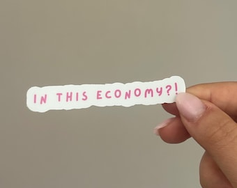 May include: A white sticker with the phrase "IN THIS ECONOMY?!" in pink handwritten font. The sticker has a torn edge and is held by a hand against a neutral background.