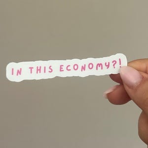 May include: A white sticker with a pink handwritten text that reads "IN THIS ECONOMY?!" The sticker has a rough, uneven edge. The sticker is held by a person with light skin and pink nail polish.