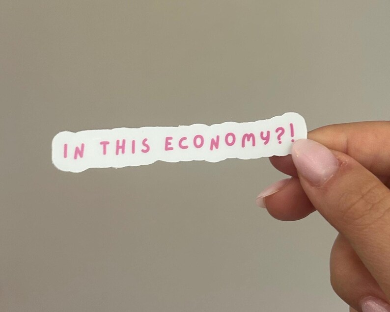 May include: A white sticker with a pink handwritten text that reads "IN THIS ECONOMY?!" The sticker has a rough, uneven edge. The sticker is held by a person with light skin and pink nail polish.