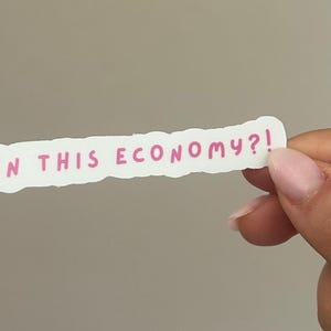 May include: A white sticker with a torn edge, featuring the phrase "IN THIS ECONOMY?!" in pink lettering. The sticker is held by a hand against a neutral gray background.
