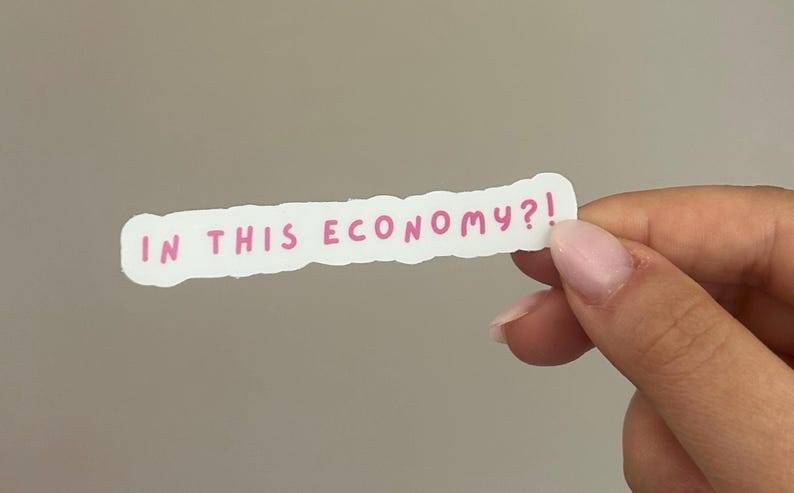 May include: A white sticker with a torn edge, featuring the phrase "IN THIS ECONOMY?!" in pink lettering. The sticker is held by a hand against a neutral gray background.