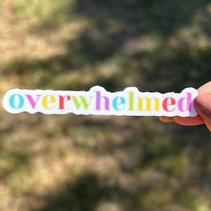 Op de afbeelding: Een witte sticker met het woord "overwhelmed" erop geschreven in regenboogkleuren. De sticker wordt vastgehouden door een hand.