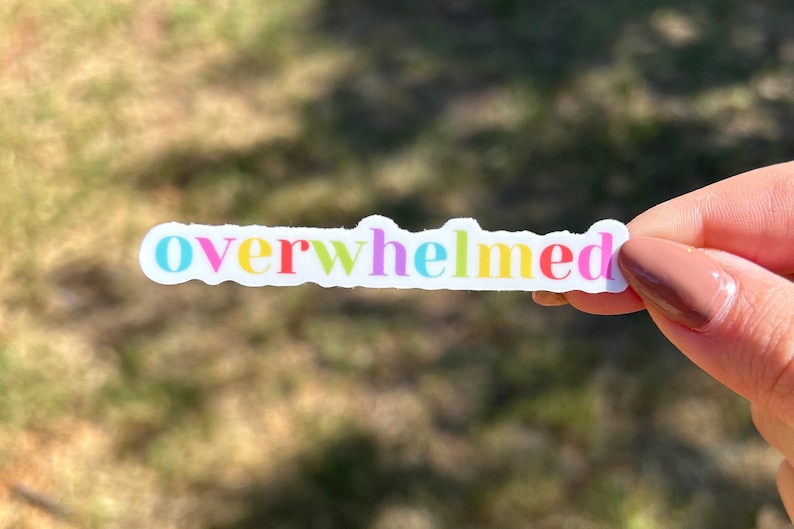 Op de afbeelding: Een witte sticker met het woord "overwhelmed" erop geschreven in regenboogkleuren. De sticker wordt vastgehouden door een hand.