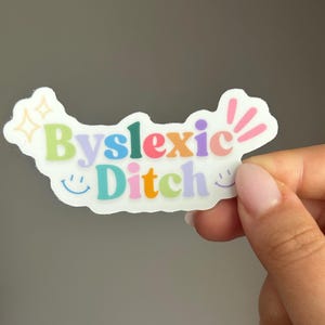Puede incluir: Una pegatina blanca con la frase "Byslexic Ditch" en letras de burbujas coloridas. La pegatina tiene un contorno ondulado, con pequeñas estrellas y rayos rosas en los bordes, y caras sonrientes en la parte inferior.
