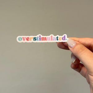 May include: A sticker with the word "overstimulated" in colorful letters. The sticker is held by a hand with manicured nails against a gray background. The text is in a playful font with each letter a different color.
