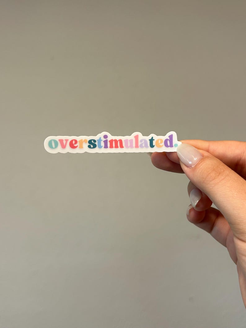 May include: A sticker with the word "overstimulated" in colorful letters. The sticker is held by a hand with manicured nails against a gray background. The text is in a playful font with each letter a different color.