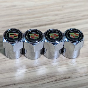 May include: Four chrome-finished tire valve stem caps. Each cap features a black emblem with the Cadillac crest in red, green, and gold, surrounded by a laurel wreath. The caps are hexagonal in shape.
