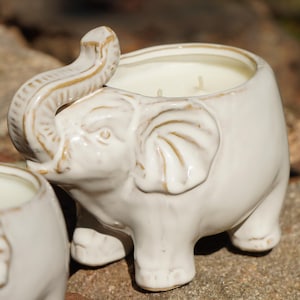 May include: Two white ceramic elephant candle holders with a white candle inside each. The candle holders are sitting on a rock.