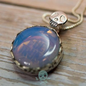 May include: Silver-toned pendant necklace featuring a large, round, opalescent stone with a skull accent. The stone has a blue and white color gradient. The necklace is on a silver chain. The background is a wooden surface.