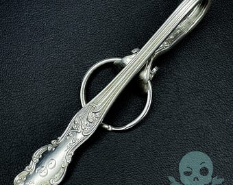 May include: A silver-plated spoon keychain with an ornate design and a ring attached. The spoon is bent into a hook shape and has a decorative handle.