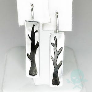 May include: A pair of silver dangle earrings featuring a black tree branch design. The earrings are rectangular in shape and have a simple, minimalist style.