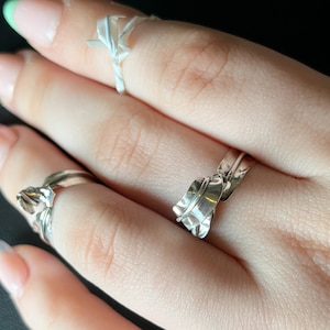 May include: Two silver rings on a hand. One ring is a simple band, the other is a more intricate design with a textured, leaf-like shape.