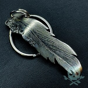 以下が含まれることがあります： 複雑なディテールと円形のリングが付いた銀製の羽根の形をしたキーホルダー。羽根はテクスチャ加工された表面と上部に詳細なデザインが施されています。
