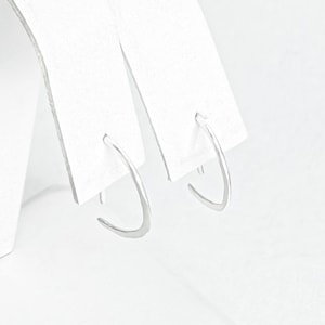 May include: A pair of silver hoop earrings with a slightly curved shape. The earrings are displayed on a white card.