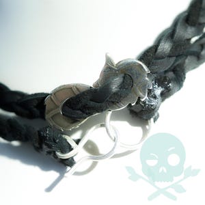 May include: Close-up of a black braided leather bracelet with a silver-colored clasp. The clasp features a decorative leaf design. The bracelet is secured with a silver-colored hook and eye closure. The background is white.