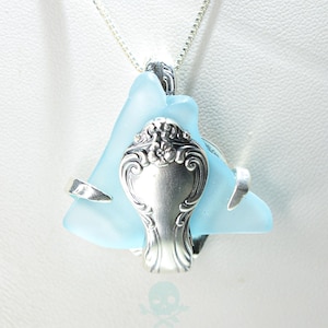 Puede incluir: Un collar con colgante único que presenta un trozo de vidrio marino azul claro con un diseño decorativo de mango de cuchara plateada. El colgante cuelga de una delicada cadena de plata, creando una pieza de joyería única.