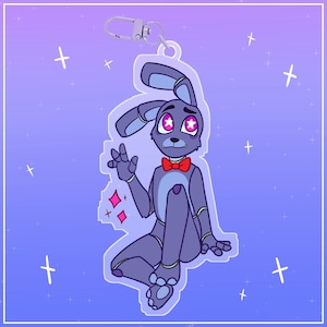 May include: A blue bunny rabbit character with a red bow tie and pink star-shaped eyes. The character is sitting with its right arm raised in a wave. The character is on a clear background with a silver keychain attached.