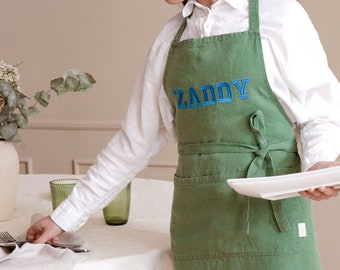 May include: A green linen apron with the word "ZADDY" embroidered in blue on the front. The apron has two pockets and a tie at the waist.