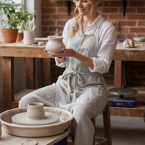Puede incluir: Una mujer con un delantal y pantalones de color gris claro sostiene un jarrón de cerámica en un estudio de cerámica. Un torno de alfarero con una olla parcialmente formada está en primer plano. El estudio tiene una pared de ladrillo y mesas de madera con artículos de cerámica.