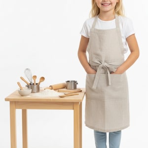 May include: A beige linen apron with a front pocket and tie waist. A young person wearing the apron stands next to a small wooden table with baking tools and flour. The table includes a rolling pin, whisk, and small bowls.