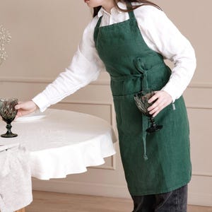 May include: A dark forest green apron featuring a bib front, adjustable neck strap, and a waist tie that secures in a bow. The apron includes two large front pockets and is constructed from a textured fabric, worn over a white long-sleeve shirt.