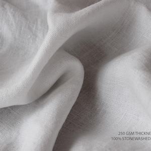 K&ouml;nnte beinhalten: Nahaufnahme eines wei&szlig;en, steinwaschgewaschenen Leinenstoffes. Der Stoff hat eine strukturierte, gewebte Optik. Der Text "250 GSM THICKNESS 100% STONEWASHED LINEN" ist in der rechten unteren Ecke des Bildes sichtbar.