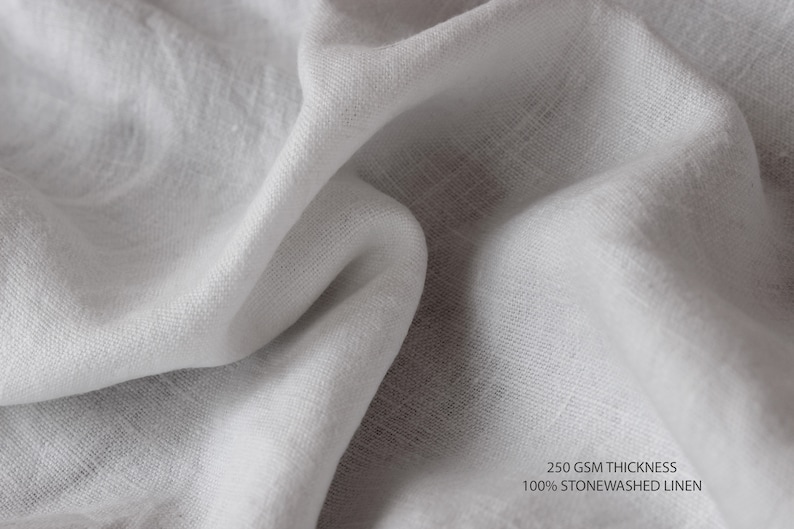 K&ouml;nnte beinhalten: Nahaufnahme eines wei&szlig;en, steinwaschgewaschenen Leinenstoffes. Der Stoff hat eine strukturierte, gewebte Optik. Der Text "250 GSM THICKNESS 100% STONEWASHED LINEN" ist in der rechten unteren Ecke des Bildes sichtbar.