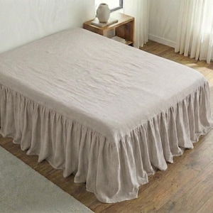 Può includere: Un letto beige con una gonna a balze. Il letto è coperto da un lenzuolo beige abbinato. Il letto è in una stanza con pavimento in legno e un piccolo comodino in legno.